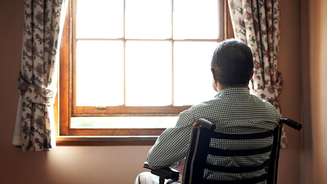Homem idoso em cadeira de rodas visto de costas olhando pela janela