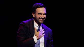 Zohran Mamdani comemora a vitória, sorridente, com uma das mãos no peito. Ele está de terno, com gravata azul-celeste, sobre fundo preto.