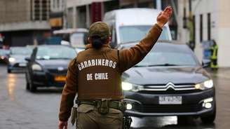Uma policial chilena vista de costas vestindo uniforme marrom com colete a prova de balas onde se lê "Carabineros de Chile". Ela tem o braço direito erguido e faz sinal para veículos na rua desviarem