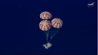 Imagem aérea mostra três paraquedas com listras brancas e vermelhas inflados amortecendo a aterrissagem da cápsula