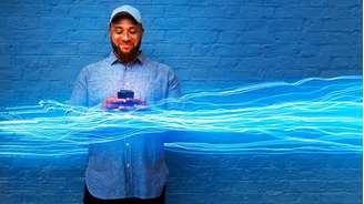 Homem sorrindo com seu celular em frente a uma parede de tijolos pintada de azul, com uma ilustração de feixes de luz azul passando por ele