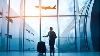 Uma mulher com bagagem está de costas para a câmera, olhando para um avião através da janela.