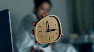 Pessoa sentada na cama mostra um despertador marcando três horas