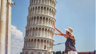 Uma mulher loira vestindo uma blusa roxa e um chapéu de sol posando para uma foto em frente à Torre Inclinada de Pisa para parecer que ela a está segurando.