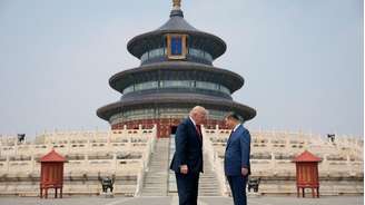 Os presidentes Trump e Xi estão em pé, frente a frente e com a cabeça levemente inclinada, diante de uma escadaria ornamentada que leva a um templo chinês decorado em tons de azul, verde, vermelho e dourado.
