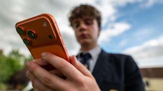 Um adolescente vestindo um uniforme escolar da Marinha, blazer, gravata e camisa branca segura um smartphone laranja à sua frente.