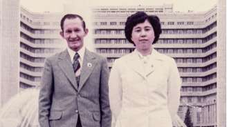 Uma fotografia antiga de Hitomi e seu marido, Charles. Eles estão em pé em frente a uma fonte e um prédio de apartamentos. Hitomi veste um blazer com botões e uma saia. Charles usa um terno cinza com uma gravata listrada. Hitomi está com os braços atrás das costas e Charles com os braços ao lado do corpo. Ambos sorriem e olham diretamente para a câmera.