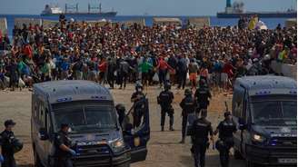 Migrantes que atravessaram do Marrocos para a Espanha retornam a uma praia após terem sido expulsos da área no dia anterior, enquanto a polícia espanhola os escolta no enclave espanhol de Ceuta, na sexta-feira, 21 de agosto de 2026.