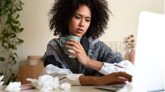 Uma mulher enrolada em um cobertor e segurando uma bebida quente sentada diante de uma mesa com um laptop. Há também lenços de papel amassados sobre a mesa