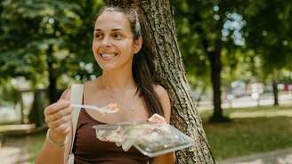 Mulher jovem comendo salada e sorrindo em frente a uma árvore.