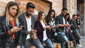 Oito jovens, lado a lado, encostados em uma parede, olhando seus telefones celulares