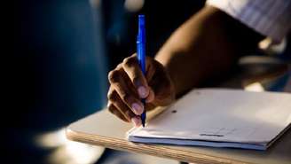 Uma mão negra segura uma caneta de tubo azul posicionada em cima de um caderno