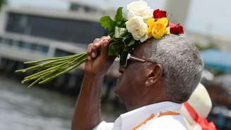 Imagem mostra homem negro, de branco, pronto para jogar flores no mar.