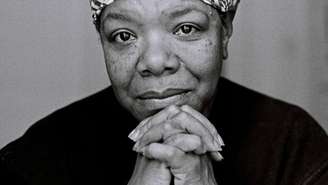 A imagem está em preto e branco e enquandra o rosto de uma mulher negra, que está com as mãos juntas abaixo do queixo