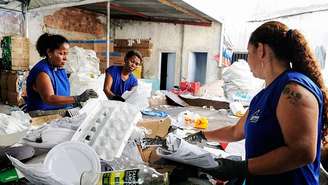 Três mulheres negras usam coletes azuis e estão catando recicláveis