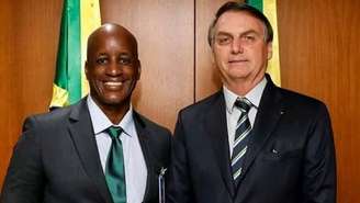 A imagem mostra Sergio Camargo, à esquerda e Jair Bolsonaro, à direita de pé e sorrindo. Eles vestem terno e gravata escuros