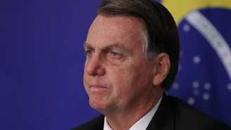 A imagem mostra o presidente Jair Bolsonaro, com o rosto em destaque. Atraz está a bandeira do Brasil
