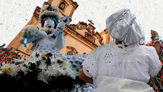 Foto mostra uma imagem de uma igreja católica ao fundo e uma mulher com vestes típicas de candomblé de costas, representando o sincretismo religioso