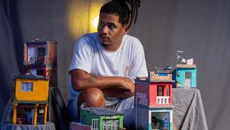 Marcelino é um homem negro que usa dreads. Ele está sentado ao centro da imagem com miniaturas de casas ao redor
