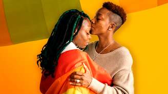 Imagem mostra duas mulheres negras envolvidas na bandeira da comunidade LGBT. Uma beija a testa da outra.