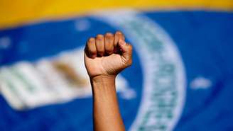 Imagem mostra a bandeira do Brasil ao fundo e a mão fechada de uma pessoa negra para cima.
