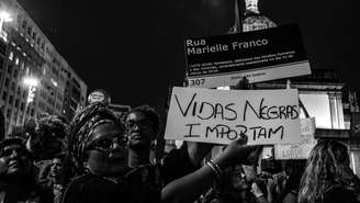 Imagem mostra mulher negra segurando cartaz contra o genocídio da população negra e em homenagem à vereadora Marielle Franco, assassinada em 2019.