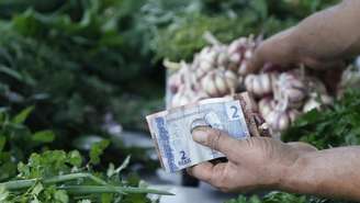 Imagem mostra mão de pessoa segurando cédulas de real, enquanto escolhe alimentos.