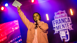 Um homem com um dos braços levantado com uma ficha na mão, e a outra mão segurando um microfone na altura da boca