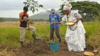 Dois homens e uma mulher em um campo aberto, de mato seco e montanhas ao fundo. A mulher usa vestimentas de religiões afro.