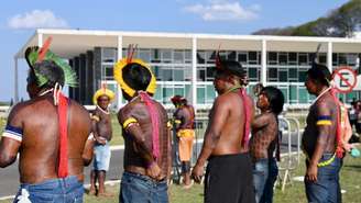 A imagem mostra sete pessoas indígenas lado a lado, sendo somente a segunda delas virada para frente e as outras estão de costas. Eles têm a pele escura, com tom avermelhado, estão sem camisa e vestem adornos tradicionais, como cocar e braceletes. No fundo, a sede do STF, onde acontece o julgamento do Marco Temporal