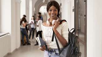 Aluna negra com mochila nas costas e livro nas mãos em uma faculdade.