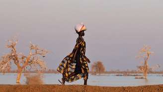 Uma mulher com um vestido preto com abacaxis estampados caminha ao longo de um dique de terra com um pacote na cabeça. Do outro lado do dique há várias árvores rodeadas de água das cheias - Bentiu, Sudão do Sul