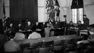 Uma mulher alemã, que acusa três soldados norte-americanos de estupro, presta depoimento diante de oficiais superiores em uma prefeitura na Alemanha. Atrás dela, há uma pintura que mostra soldados da Wehrmacht, do Volkssturm e da Juventude Hitlerista empunhando bandeiras. Esse foi o primeiro julgamento por corte marcial da história da 7ª Divisão do Exército dos Estados Unidos realizado em solo alemão.