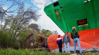 Três pessoas, vestindo jeans e jaquetas casuais, estão de pé, observando um enorme navio porta-contêineres verde que encalhou em terra firme. Uma casa parece pequena e está ao lado do navio.