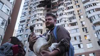Um homem carrega seu cachorro em frente a um prédio residencial de vários andares danificado após um ataque de drone russo em Kiev, Ucrânia, nesta sexta-feira