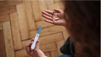 Mulher segura aparelho de teste de gravidez com uma mão e pílula na outra, olhando para o chão