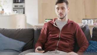 Um homem olhando para a TV com rosto de tédio e segurando um controle remoto