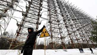Pessoa de pé em frente a uma enorme antena de radar, ao lado de uma placa com o símbolo de radiação
