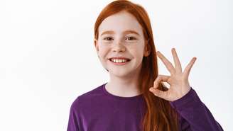 Happy redhead child, ginger girl with freckles shows okay sign in approval, smiling satisfied, looking cheerful and pleased, standing over white background.
