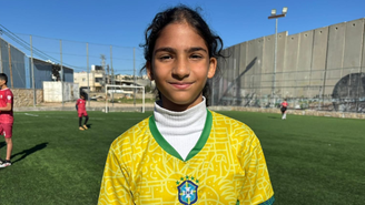 Uma menina palestina olha para a câmera. Ela está em um campo de futebol e veste a camisa da seleção brasileira