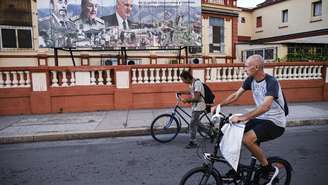 Moradores circulam de bicicleta nas ruas de Havana, em frente a um cartaz com imagens do presidente de Cuba, Miguel Díaz-Canel (à dir.), e de seus antecessores no cargo, Fidel (à esq.) e Raúl Castro, em 16 de fevereiro de 2026.