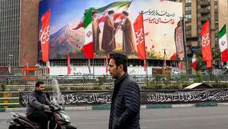 Um homem caminha em Teerã e, ao fundo, um mosaico dos líderes da Revolução Islâmica