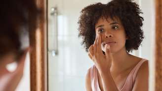 Uma jovem de cabelos escuros olha-se no espelho, tocando a pele ao redor dos olhos com uma expressão sombria