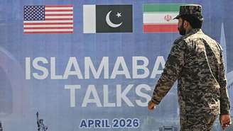 Um guarda paquistanês, vestindo um uniforme de camuflagem, passa por um outdoor sobre as negociações de paz entre os EUA e o Irã em Islamabad