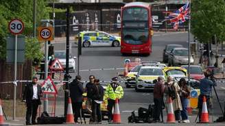 Uma cena de rua mostra membros da imprensa trabalhando perto de um cordão policial, em frente a carros da polícia e um ônibus estacionado, enquanto uma bandeira britânica tremula acima deles