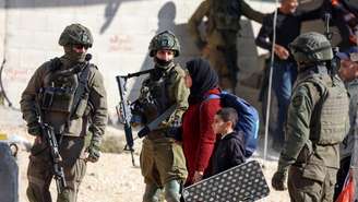 Cisjordânia: 70 crianças palestinas mortas desde o início das operações israelenses (ONU). Na foto, uma moradora palestina do campo de refugiados de Nur Shams caminha com uma criança ao lado de soldados israelenses, carregando pertences retirados de sua casa antes da demolição de edifícios residenciais no campo, perto de Tulkarem, na Cisjordânia ocupada por Israel, em 17 de dezembro de 2025.