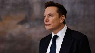 Elon Musk vestindo um terno preto, camisa branca e gravata preta olhando para longe