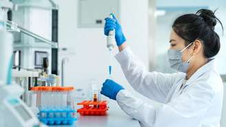 Um ambiente de laboratório com uma pessoa com um jaleco branco trabalhando em uma bancada. A pessoa usa luvas de proteção e máscara facial enquanto usa cuidadosamente uma pipeta para transferir o líquido para um pequeno tubo de ensaio. Várias prateleiras de tubos de ensaio com líquidos coloridos estão dispostas ordenadamente na superfície de trabalho em primeiro plano. Equipamentos científicos e prateleiras são visíveis ao fundo, sugerindo um espaço de trabalho limpo e organizado