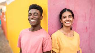Um homem e uma mulher jovens aparecem lado a lado diante de uma parede pintada com cores vibrantes. Ele veste uma camiseta rosa diante de uma parte amarela da parede, enquanto ela usa uma camiseta amarela diante de uma área rosa