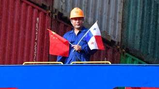 Um trabalhador vestido com macacão azul e capacete de proteção laranja segura as bandeiras da China e do Panamá